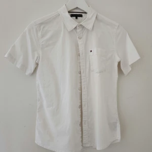 Vit kortärmad skjorta Tommy Hilfiger - Klassisk vit skjorta från Tommy Hilfiger i bomull med korta ärmar, bröstficka med liten logga och knappar framtill. Skjortan har en stilren och enkel design med normal passform och mjukt material som känns skönt mot huden.Storlek 146-152