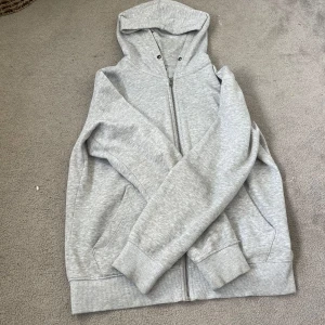 Ljusgrå hoodie med dragkedja - Enkel och stilren ljusgrå hoodie från Jack&Jones. Tröjan har långa ärmar, ribbade muddar och två fickor på sidorna. Perfekt för chill dagar eller när du vill ha något bekvämt och basic. Snörerna i luvan är tyvärr utdragna 