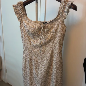 Blommig kortklänning från SHEIN - Supersöt kortklänning från SHEIN med blommigt mönster i beige och lila toner. Klänningen har volangdetaljer på axelbanden och knytning fram.