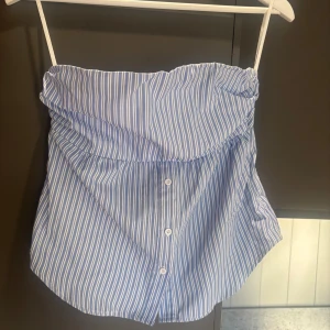 Blåvit randig tubtopp med knappar - Säljer en blå och vit randig tubtopp med knappar framtill. Toppen har bandeau-modell och är sydd i bomullstyg med klassiska skjortränder. Perfekt för en fräsch och avslappnad look i sommar.