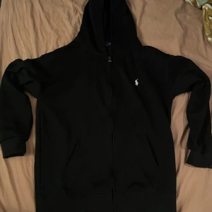 Svart hoodie från Polo Ralph Lauren - Svart hoodie från Polo Ralph Lauren i 100% bomull. Klassisk modell med huva, dragkedja framtill och känguruficka. Vit broderad logga på bröstet ger en clean och stilren look. Perfekt för chill dagar eller när du vill ha en enkel men snygg outfit.