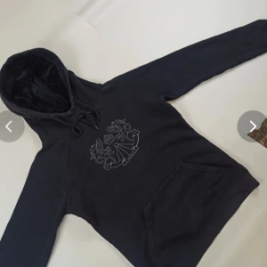 Svart hoodie med draktryck och huva - Svart hoodie med coolt tryck av två drakar och krona på bröstet. Tröjan har huva med snörning och en stor magficka framtill. Perfekt för dig som gillar streetstyle och vill ha en bekväm och snygg look.