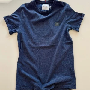 Mörkblå t-shirt från Les Deux - Snygg mörkblå t-shirt från Les Deux med klassisk passform och rund hals. T-shirten har korta ärmar och en diskret logotyp-patch på bröstet. Tillverkad i mjuk bomull som känns skön mot huden. Perfekt för en clean och stilren look.