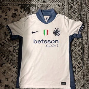 Inter Milan vit bortatröja Nike - Inter Milan bortatröja i vitt med blå detaljer och klassisk krage. Tryck med klubbmärke, Nike-logga och sponsor på bröstet. Materialet är lätt och andas, perfekt för match eller träning. Snygga blå ränder på ärmsluten och logga på ryggen.