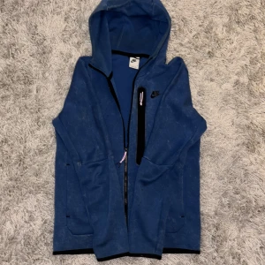 Blå hoodie från Nike med dragkedja - Säljer en blå hoodie från Nike i storlek S. Tröjan har hel dragkedja framtill, huva och svarta detaljer vid dragkedjan och loggan. Perfekt för chill dagar eller när du vill ha en sportig look. Mjuk och skön med fickor på sidorna. Hör av er om ni vill ha fler bilder! Pris kan diskuteras.