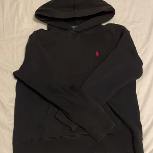 Svart hoodie från Polo Ralph Lauren - Svart hoodie från Polo Ralph Lauren med klassisk röd broderad logga på bröstet. Tröjan har huva, känguruficka och långa ärmar. Perfekt för en chill och stilren look.