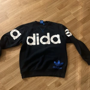 Mörkblå sweatshirt från Adidas - Mörkblå sweatshirt från Adidas med stor vit logga över bröstet och ärmarna samt blå Adidas-tryck nertill. Klassisk rund halsringning och långa ärmar. Perfekt för en sportig och avslappnad stil.