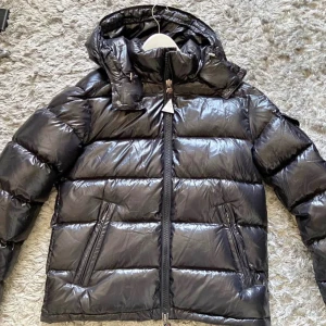 Moncler Maya  - Snygg svart dunjacka från Moncler med glansig finish och klassisk puffer-design. Jackan har huva, två snedställda fickor med dragkedja och Moncler-logga på ärmen. Perfekt för kalla dagar och riktigt street. Stängs med dragkedja hela vägen upp, storlek 2