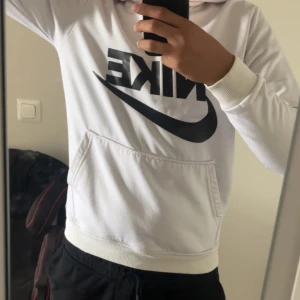 Vit hoodie från Nike med svart logga - Säljer en vit hoodie från Nike med stor svart logga och swoosh på bröstet. Tröjan har huva, känguruficka och ribbade muddar vid ärmslut och midja. Perfekt för dig som gillar sportig och clean stil.