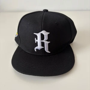 Svart baseballa keps - Snygg svart keps med platt skärm och stor vit broderad R på framsidan. Justerbar snapback baktill och färgglada broderade detaljer på sidan. Perfekt för dig som gillar streetstyle och vill sticka ut med en unik accessoar. Helt ny.