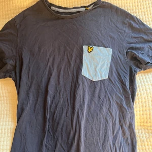 Blå t-shirt med ficka från Lyle & Scott - Snygg mörkblå t-shirt från Lyle & Scott med ljusblå bröstficka och klassisk gul logga. T-shirten är i mjuk bomull och har en normal passform med korta ärmar. Perfekt för en chill och stilren look.