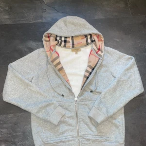 Grå Burberry hoodie med rutig huva - Snygg grå hoodie från Burberry med klassisk rutig insida i huvan och broderad logga på bröstet. Tröjan har dragkedja, snörning i huvan och är tillverkad i mjuk bomull. Perfekt för dig som gillar stilrena detaljer och exklusiva märken.