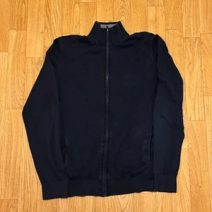 Mörkblå kofta från Pier One - Mörkblå kofta från Pier One med hel dragkedja framtill och ribbad ståkrage i grått. Tröjan är långärmad och har två fickor på sidorna. Tillverkad i mjuk bomull, perfekt för lager-på-lager under kyliga dagar.