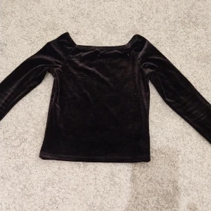 Svart sammetstopp från Kappahl - Mörk, svart långärmad topp i mjuk sammet från Kappahl. Toppen har en rak, enkel passform och bredare halsringning som ger en stilren look. Perfekt för dig som gillar en klassisk men ändå trendig vibe.