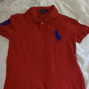 Röd pikétröja Polo Ralph Lauren S - Röd pikétröja från Polo Ralph Lauren i slim fit med klassisk krage och knappar. Stor blå broderad logga på bröstet och blå siffra 3 på ärmen. Tillverkad i bomull, perfekt för en sportig och stilren look.