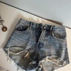 Blå jeansshorts med fransar från Zara - Snygga blå jeansshorts från Zara med hög midja och råa, fransiga kanter. De har slitna detaljer framtill, klassiska fem fickor och knappstängning. Använda 2 ggr under sommaren, inga defekter och nyskick! 