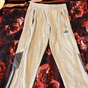 Grå Adidas mjukisbyxor med dragkedjor - Grå mjukisbyxor från Adidas med klassiska tre ränder längs benen och logga på låret. Byxorna har dragkedjor på fickorna, snörning i midjan och detaljer i mörkare grått vid knäna. 