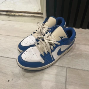 Nike Air Jordan 1 Low blå/vit 42.5 - Nike Air Jordan 1 Low i blått och vitt skinn med klassisk Air Jordan-logga på hälen. Snygg kontrast mellan blåa och vita paneler, perforerad tå och vit sula. Låga sneakers med snörning och ikonisk swoosh på sidan.