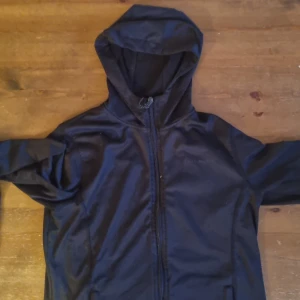 Svart sportjacka från True North - Svart sportjacka från True North med huva och hel dragkedja framtill. Jackan har mesh/fleecefoder på insidan för extra komfort och andningsförmåga. Perfekt för blåsiga dagar och enkel att slänga på sig när vädret svänger.