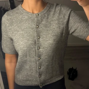 Grå stickad kofta med kort ärm - Superfin grå stickad kofta med korta ärmar och snygga guldfärgade knappar framtill. Mjuk och skön känsla, enkel att matcha med både jeans och kjol. Säljer då jag aldrig använt den! Storlek S