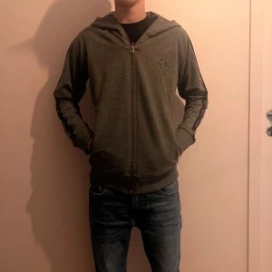 Grå hoodie från Calvin Klein - Mörkgrå hoodie från Calvin Klein med dragkedja framtill och huva. Diskret CK-logga på bröstet och Calvin Klein-tryck längs ärmarna. Tröjan har långa ärmar och två fickor med dragkedja. Perfekt för en chill och stilren look.
