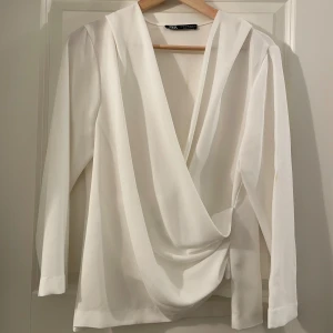 Vit omlottblus från Zara - Säljer en stilren vit omlottblus från Zara i storlek M. Blusen har lång ärm, djup v-ringning och snyggt draperad front. Perfekt att styla med jeans eller kjol för en clean look.