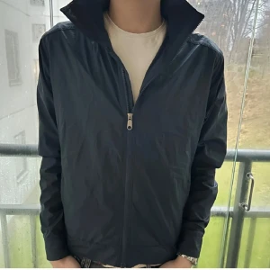 Svart vindjacka Peak Performance M - Snygg svart vindjacka från Peak Performance i storlek M. Jackan har hög krage, dragkedja framtill och är tillverkad i lätt polyester. Perfekt för dig som gillar stilren och sportig look. Passformen är normal och jackan är helfärgad utan mönster.