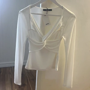 Vit omlottopp från Bik Bok - Säljer en stilren vit omlottopp från Bik Bok med lång ärm och snygg v-ringning. Toppen har en figurnära passform och är tillverkad i ett mjukt, stretchigt material som sitter skönt på kroppen. Perfekt för dig som gillar trendiga och enkla plagg. (Oanvända!)