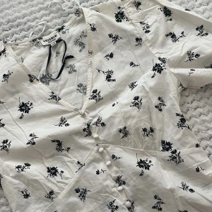 Vit blommig blus från Vintage Stories - Supersöt vit blus från Vintage Stories med svart blommigt mönster, v-ringning och knappar framtill. Korta ärmar och lätt, luftig känsla – perfekt för varma dagar. Snygg passform och romantisk stil som sticker ut.