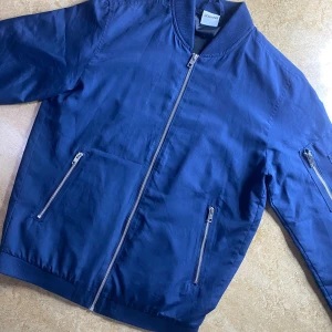 Blå bomberjacka från Jack & Jones - Snygg mörkblå bomberjacka från Jack & Jones med klassisk ribbad krage och muddar. Jackan har dragkedja framtill, två fickor med dragkedja och en ärmficka. Perfekt för dig som vill ha en stilren och enkel look.