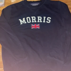 Mörkblå sweatshirt från Morris - Mörkblå sweatshirt från Morris med broderad logga och brittisk flagga på bröstet. Klassisk rund halsringning och långa ärmar. Tillverkad i mjuk bomull för en skön känsla. Perfekt för en stilren och avslappnad look.