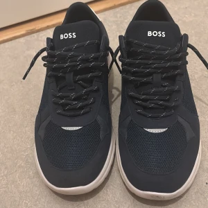 Svarta sneakers från BOSS - Snygga svarta sneakers från BOSS med vit sula och reflekterande detaljer. Ovandelen är i mesh och syntet, vilket ger en sportig vibe. Skorna har snörning med svarta och vita skosnören samt rund tå. Perfekta för dig som gillar stilrena och moderna sneakers. Dens kartong kommer med den. Tveka inte att ge era pris förslag. Skorna har används 2 gånger. 