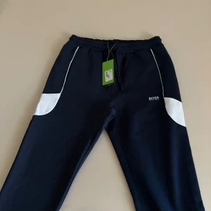 Svarta mjukisbyxor från Boss - Snygga svarta mjukisbyxor från Boss med vita detaljer längs sidorna. Byxorna har en elastisk midja och en diskret logga på framsidan. Perfekta för en avslappnad stil.