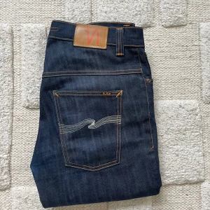 Mörkblå jeans från Nudie Jeans - Säljer ett par mörkblå jeans från Nudie Jeans med klassisk femficksdesign och kontrastsömmar. Jeansen har raka ben och snygga broderade bakfickor med vågigt mönster. Tillverkade i slitstark denim av bomull. Perfekta för dig som gillar stilrena och tidlösa jeans.
