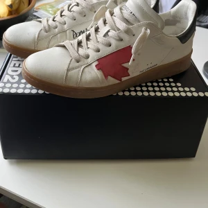 Dsquared2 Boxer sneakers vit/röd strl 42 - Snygga Dsquared2 Boxer sneakers i vitt kalvskinn med röd lönnlöv-detalj på sidan och svart häl. Klassisk snörning och gummisula. Unik print och coola handskrivna detaljer på sidan. Perfekta för dig som vill sticka ut med stil. Köpta på Johnells i Norrköping. Allt og finns, box, shoebag, kvitto.