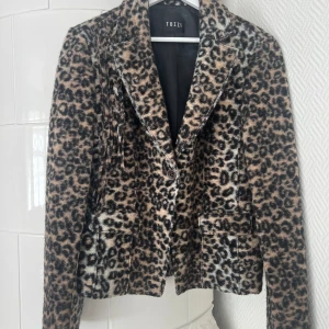 Leopardmönstrad kavaj från Tuzzi - Snygg leopardmönstrad kavaj från Tuzzi i brun, beige och svart. Kavajen har klassisk krage, knäppning framtill och två fickor. Materialet är 80% ull och 20% polyester, vilket ger en lyxig och mjuk känsla. Perfekt för dig som vill sticka ut.