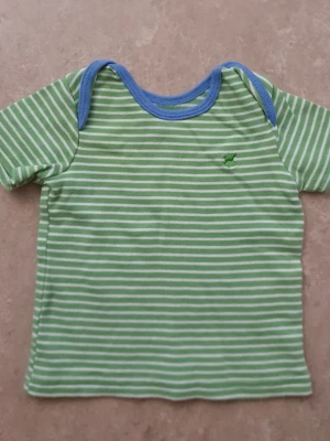 T-shirt - Säljer en grön och vit randig t-shirt med blå kant runt halsen. T-shirten har korta ärmar och ett litet broderat motiv på bröstet. Perfekt för dig som gillar färgglada och lekfulla plagg.