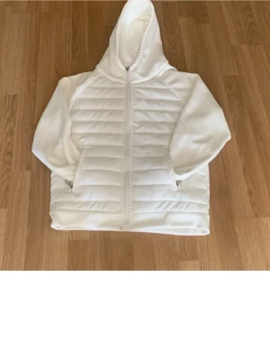 Vit quiltad pufferjacka med huva - Säljer en vit pufferjacka från JLAI med quiltad framsida och dragkedja hela vägen. Jackan har huva, långa ärmar i mjukt material och två fickor med dragkedja. Perfekt för dig som vill ha en clean och enkel look.