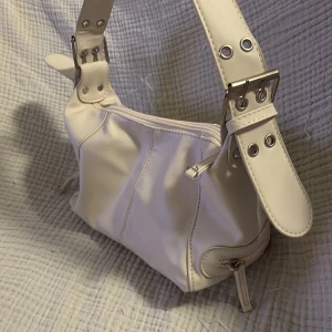 Krämvit axelväska med detaljer - Säljer en krämvit axelväska i syntetmaterial med coola metallspännen och nitar på axelbandet. Väskan har dragkedja upptill och extra dragkedjedetaljer på sidorna. Perfekt storlek för det viktigaste och ger en trendig vibe till din outfit.