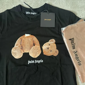 Svart t-shirt från Palm Angels med björntryck - Svart t-shirt från Palm Angels med stort tryck av en nallebjörn på framsidan och vit Palm Angels-logga på ryggen. Klassisk rund halsringning och korta ärmar. Perfekt statement-plagg för streetwear-stilen.