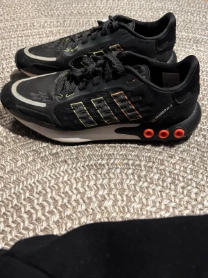 Adidas LA Trainer III S svarta sneakers strl 40 - Svarta Adidas LA Trainer III S sneakers med klassiska tre ränder i neonfärger på sidan och orange detaljer vid hälen. Skorna har snörning, platt sula och meshpartier framtill för extra ventilation. Snygg och modern design som sticker ut.
