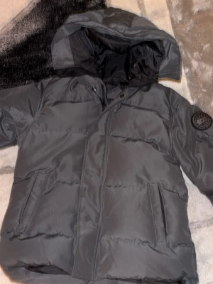 Grå pufferjacka från Canada Goose - Grå pufferjacka från Canada Goose med huva och emblem på ärmen. Jackan har quiltad design, dragkedja framtill och två sidofickor. Perfekt för kalla dagar och har en clean, stilren look som funkar till allt. Materialet är polyester och dunfyllning.