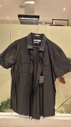Grå kortärmad skjorta Calvin Klein - Snygg grå skjorta från Calvin Klein i storlek L. Skjortan har korta ärmar, två bröstfickor med lock och knappar framtill. Tillverkad i ett lätt och bekvämt material. Helt ny med prislapp