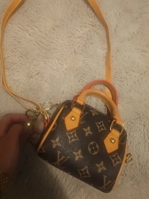  monogram axelväska - Säljer en liten och snygg axelväska med klassiskt monogrammönster i brunt och beige. Väskan har både handtag och axelrem i ljusbrunt skinn, samt guldfärgade detaljer och dragkedja. Insidan är röd med svarta ränder. Perfekt för dig som gillar lyxiga accessoarer.