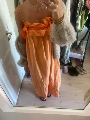 Orange långklänning med volang - Säljer en orange långklänning med smala axelband och volangdetalj upptill. Klänningen har en loose passform och är tillverkad i ett lätt bomullstyg som känns luftigt. Perfekt för varma dagar och har en snygg, enkel design med romantisk touch.