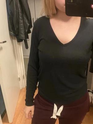 Svart ribbad långärmad topp - Säljer en svart långärmad topp med v-ringning. Toppen är ribbad och har en slim passform som sitter snyggt mot kroppen. Perfekt att matcha med jeans eller kjol för en enkel och stilren look.