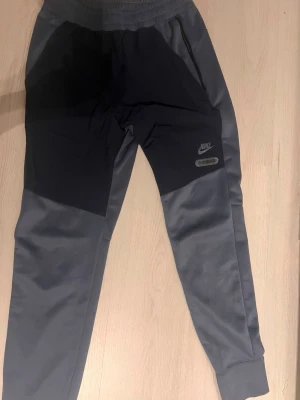 Nike Air Max mjukisbyxor grå/svart M - Nike Air Max mjukisbyxor i storlek M, med coolt tryck på benet och midjan. Byxorna är grå och svarta med elastisk midja och muddar vid benslut. Tillverkade i polyester för en skön och sportig känsla. Perfekta för chill eller träning.