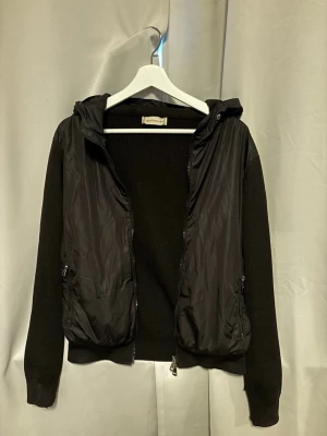Moncler cardigan black label - Svart sällsynt jacka från Moncler black label med stickade ärmar och rygg i ull, samt glansig front och huva i polyester. Jackan har dragkedja framtill och två sidofickor med dragkedja. Snyggt Moncler-märke på ärmen och ribbade muddar. Perfekt för dig som gillar stilren och sportig look. Storlek m men liten i storlek så skulle säga att den är en S. Hör av dig vid fler frågor
