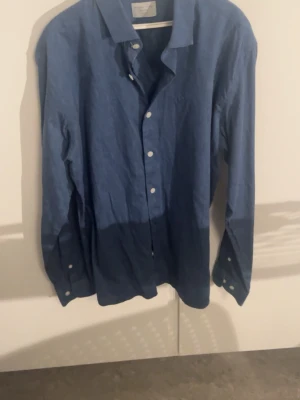 Selected Homme skjorta blå - Snygg blå skjorta från Selected Homme, modell Identity slim fit. Tillverkad i linne med klassisk krage och vita knappar framtill. Långärmad och stilren design som passar perfekt till jeans eller chinos. Skjortan har en mörkare blå ton nedtill för en cool effekt.