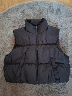 Svart dunväst från H&M Divided M - Säljer en svart dunväst från H&M Divided i storlek M. Västen har hög krage, två fickor med knappstängning och dragkedja framtill. Den är quiltad och har en loose passform, perfekt att slänga över en hoodie eller tröja när det är kyligt ute.